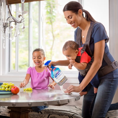 Mom cleaning table with PURELL Multi Surface Disinfectant Spray while infant strapped to her chest and daughter sitting at the table