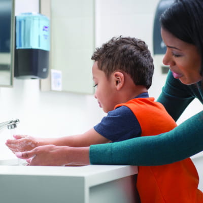 Mom helping young son wash his hands at a sink with PURELL soap dispenser nearby